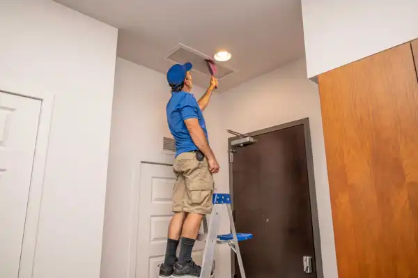 A person in a blue shirt and khaki shorts stands on a ladder, providing an air duct cleaning service as they meticulously clean an air vent on the ceiling with a brush.