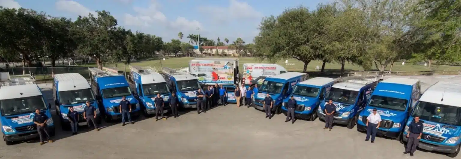 A row of nine blue service vans with people standing by them in a semi-circle, set in an outdoor area with trees and a grassy field in the background.